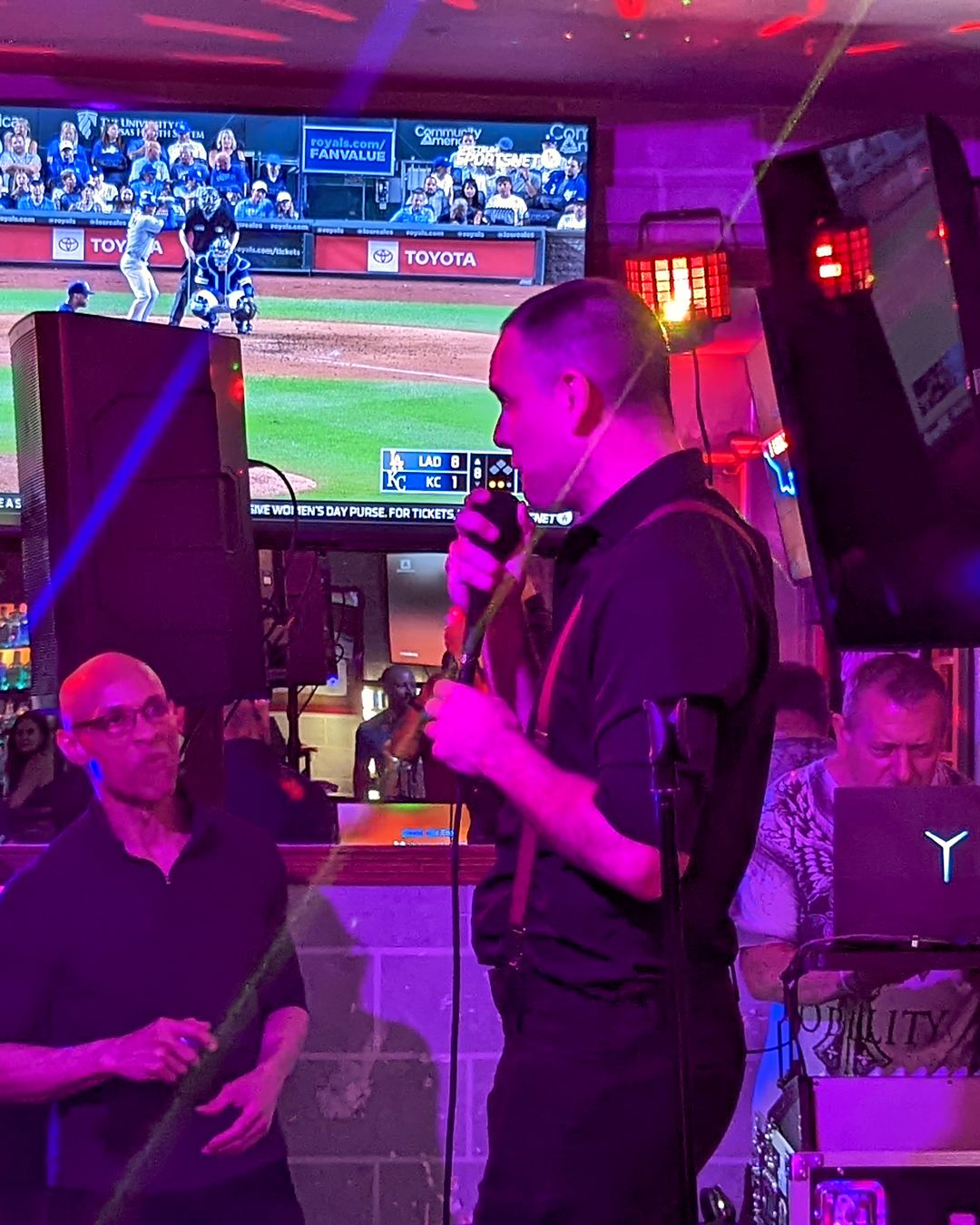 Karaoke night at Standard Station: a singer mid-song in purple light, the host at his laptop beside him, a baseball game on the big screen behind them