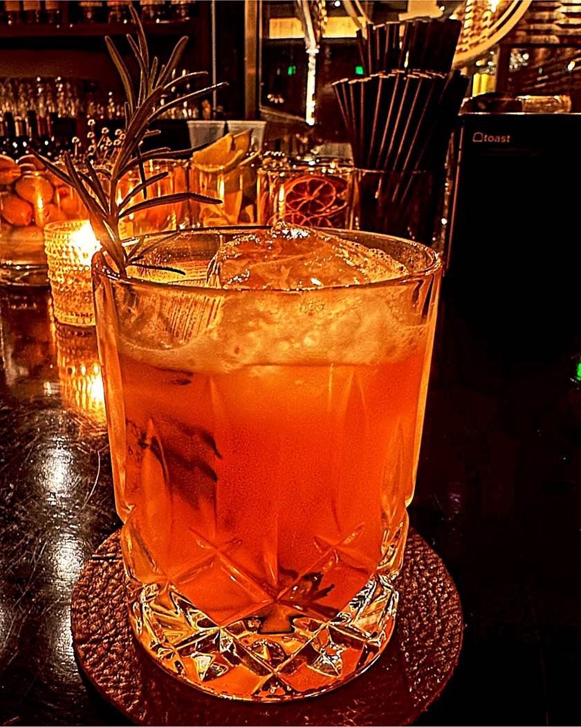 An amber cocktail in a cut crystal glass with a rosemary sprig, on the bar top by candlelight