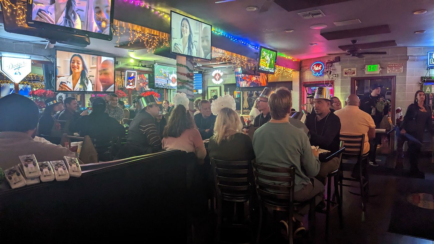 New Year's Eve crowd at Standard Station, guests in party hats under string lights, televisions over the bar